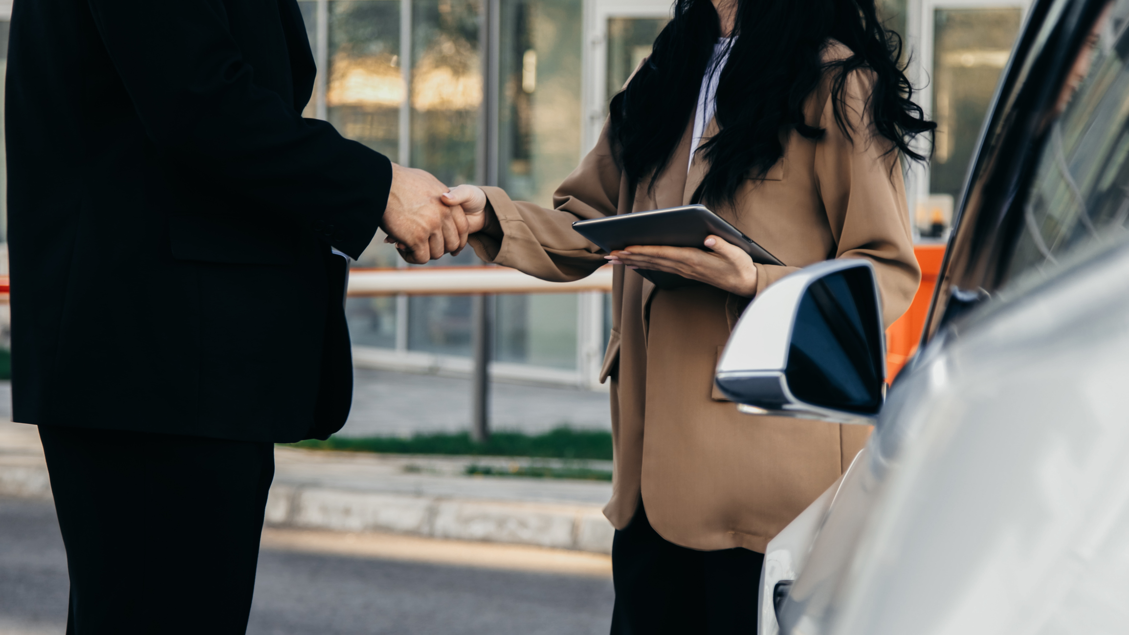 A businesswoman shakes hands with a businessman while their electric car charges in a parking lot